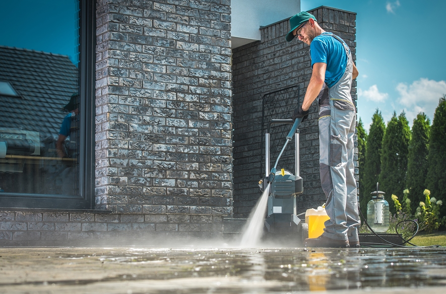 Pressure Washer Cleaning In Front Of The House. Caucasian Men In Pressure Washer Cleaning in Front of the House. Caucasian Men in His 30s Washing Concrete Bricks Driveway in Sunny Summer Day. Cleaning Around the House Concept.